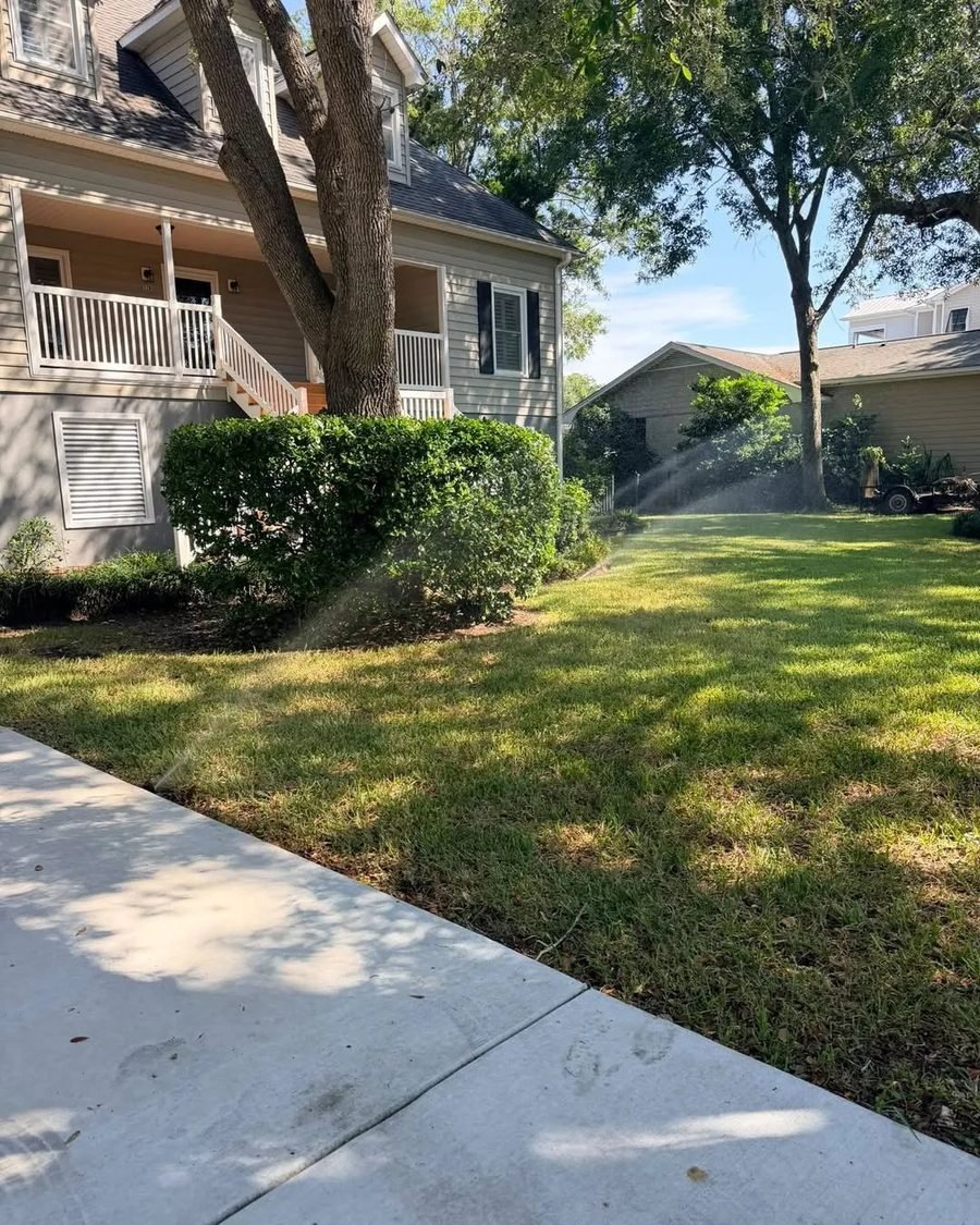 Sprinklers running across a green lawn beside a home with mature oak trees